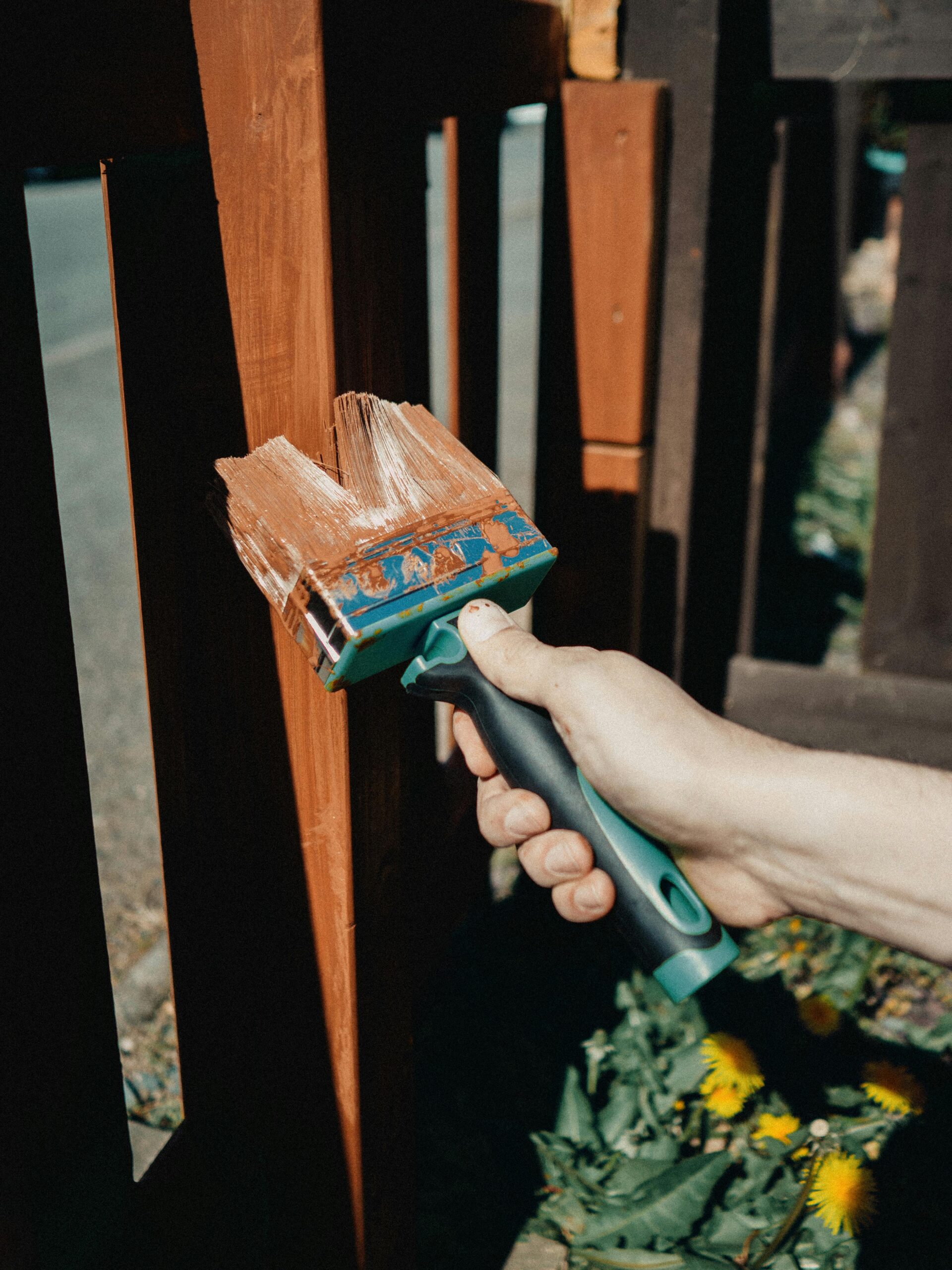 Services A close-up of a hand painting a wooden fence with a brush outdoors, surrounded by greenery.